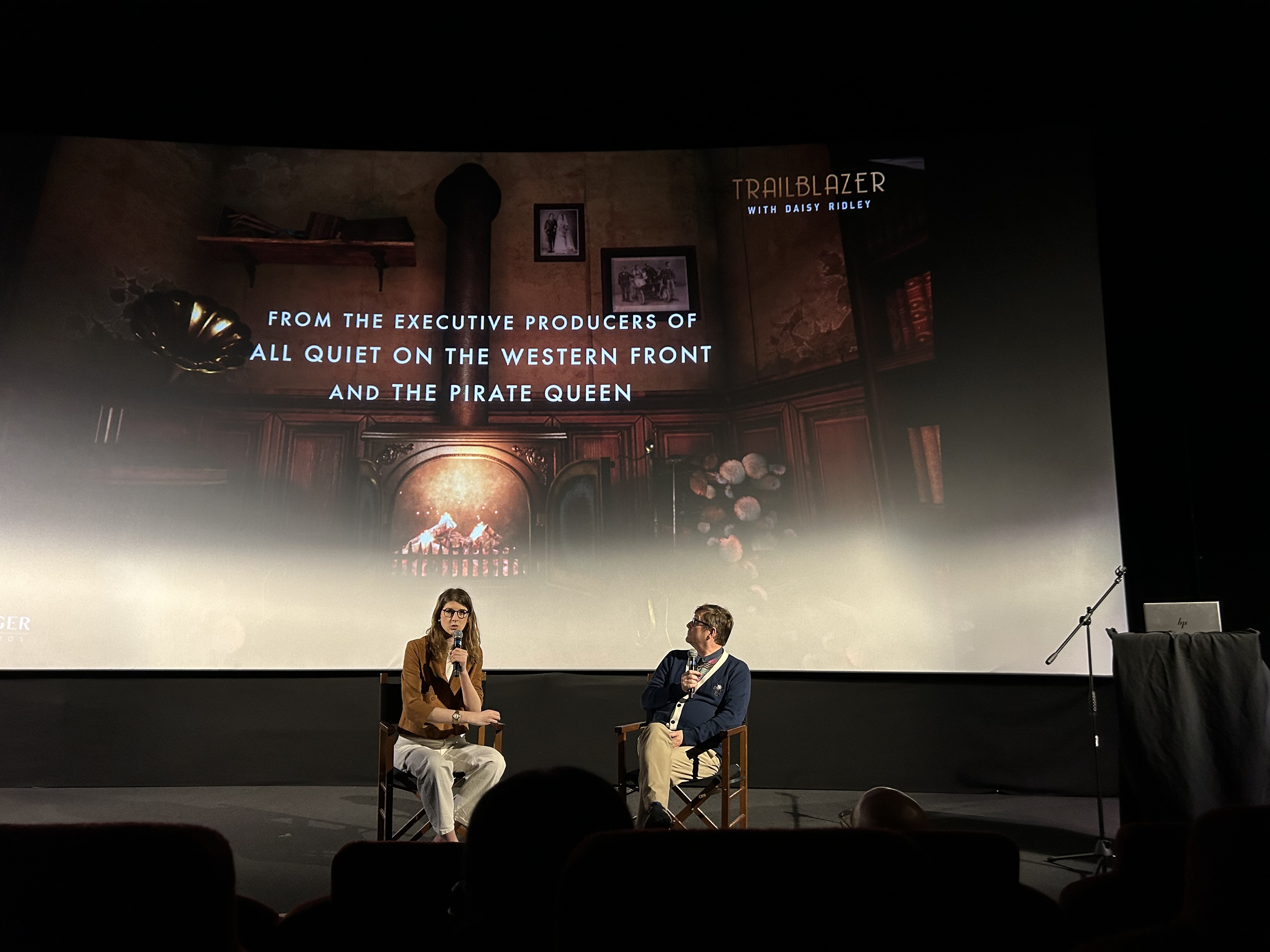 Eloise Singer and Dave Ranyard sit in front of a cinema screen with microphones, giving a presentation to an audience.