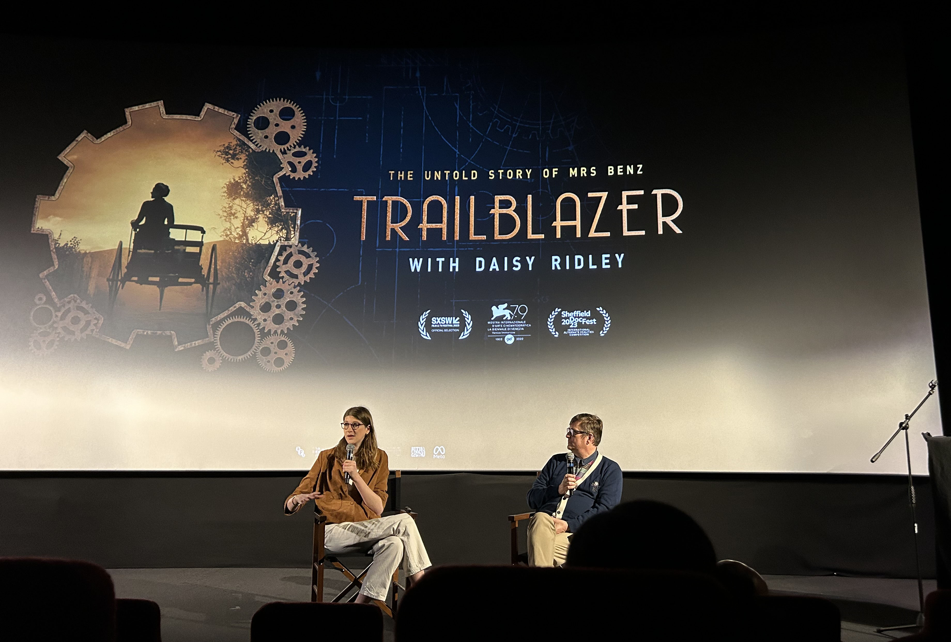 Eloise Singer and Dave Ranyard sit in director's chairs with microphones in hand, talking in front of a large cinema screen. The screen has a graphic for the game 'Trailblazer: The Untold Story of Bertha Benz'.